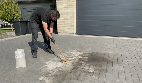 Õliplekkide eemaldamine sillutiselt, meister nühib harjaga halli betoonkivi tumedat plekki garaažiukse ees, kõrval valge kott, värske õlipleki peale soovitame enne meistri saabumist puistata imavat graanulit.