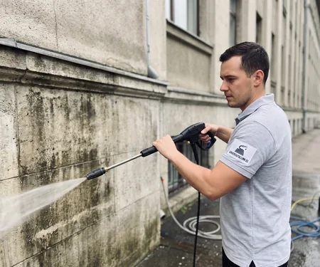 Meister peseb vana mitmekorruselise hoone määrdunud kivisoklit, karniiside alumist serva pestakse nurga all painutatud lantsiga.