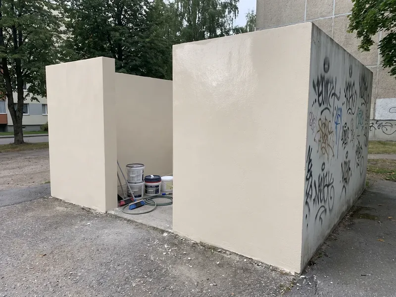Väike krohvitud prügimaja kortermajade vahel, kaks seina on puhtad ja kolmandal on veel mustad pihustivärvi tägid, sees seisavad värvipotid, rull ja voolik, prügikonteinerid veeretatakse pesu ajaks prügimajast välja ja pärast kaitsekatte kuivamist tagasi.